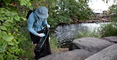 A person wearing a blue shirt and hat holding a trash bag and placing in waste picked up from the river in the background. 